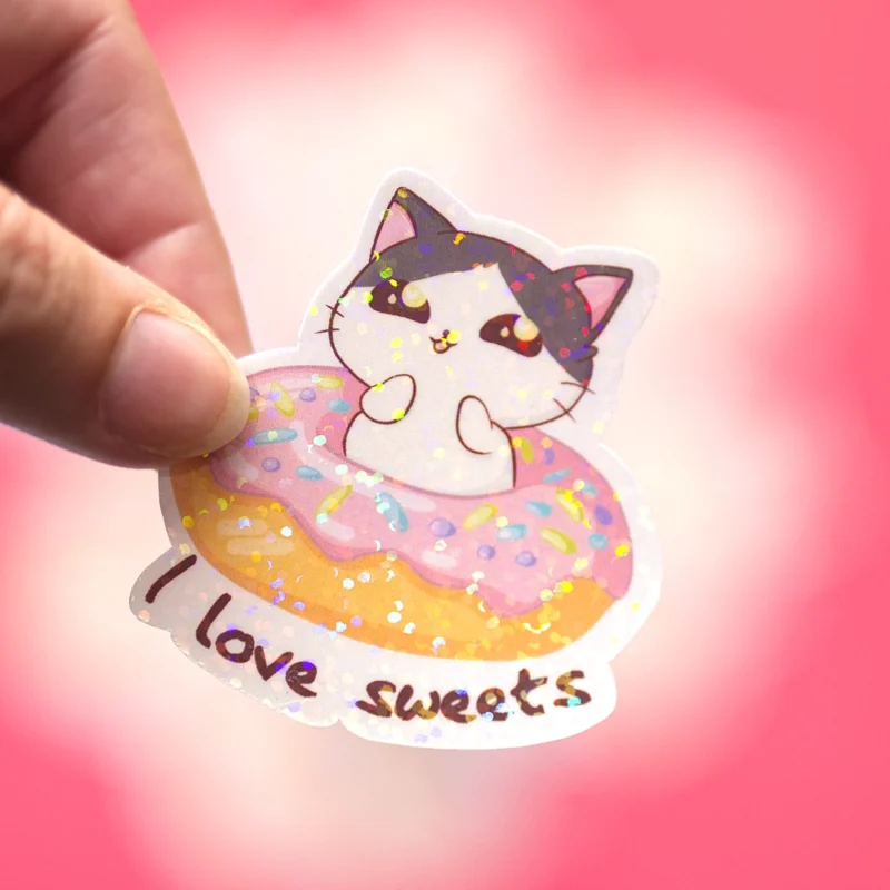 A hand holding a sticker that says I love sweets, the sticker is pink with sparkles, a cat is sitting in a dougnout.