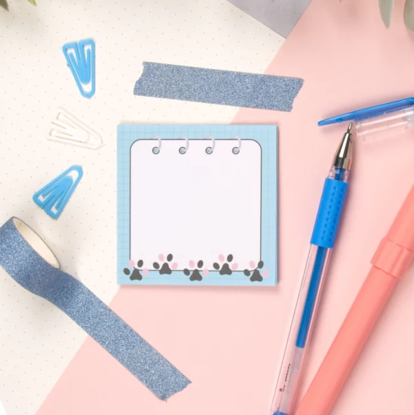 A top down picture of a sticky note pad with stationery accesories. The pad has cat paws on it.