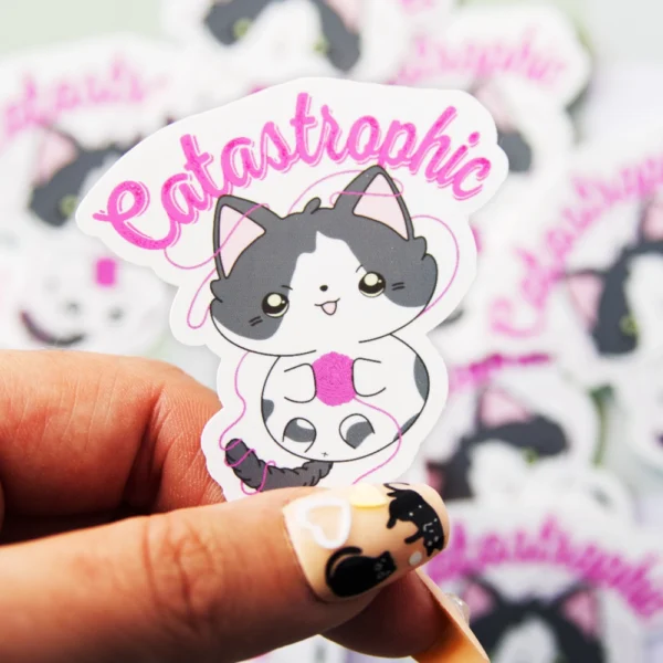 A hand holding a sticker of a cat playing with a yarn ball with the text catastrophic above it.