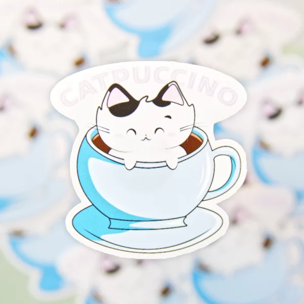 A sticker of a cat sitting in a coffee cup with the words catpuccino over it.