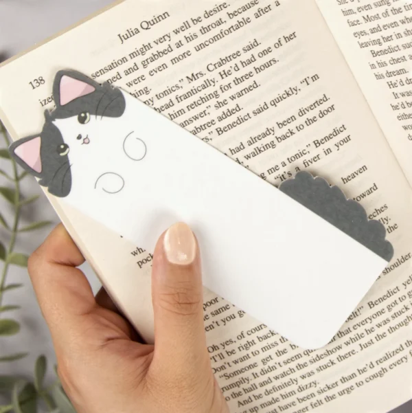 A female hand holding a bookmark on a book. Bookmark is in the shape of a long cute and happy cat.