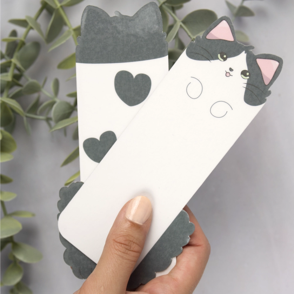 A female hand holding two bookmarks shaped like a cat. One bookmark has hearts shaped on its back.