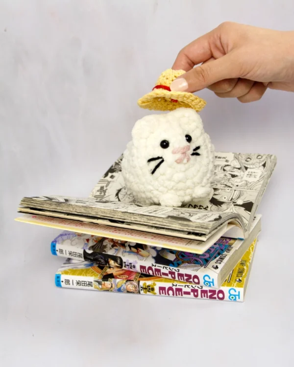 A white crochet cat sitting on top of a couple of one piece mangas. A hand is placing a crochet straw hat on the cat.