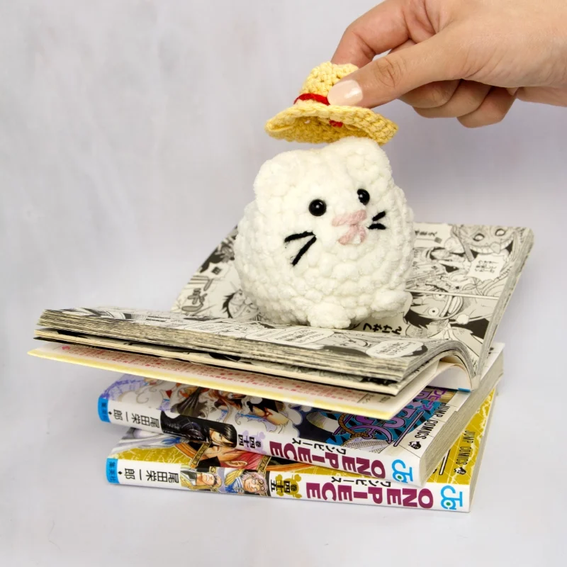 A white crochet cat sitting on top of a couple of one piece mangas. A hand is placing a crochet straw hat on the cat.