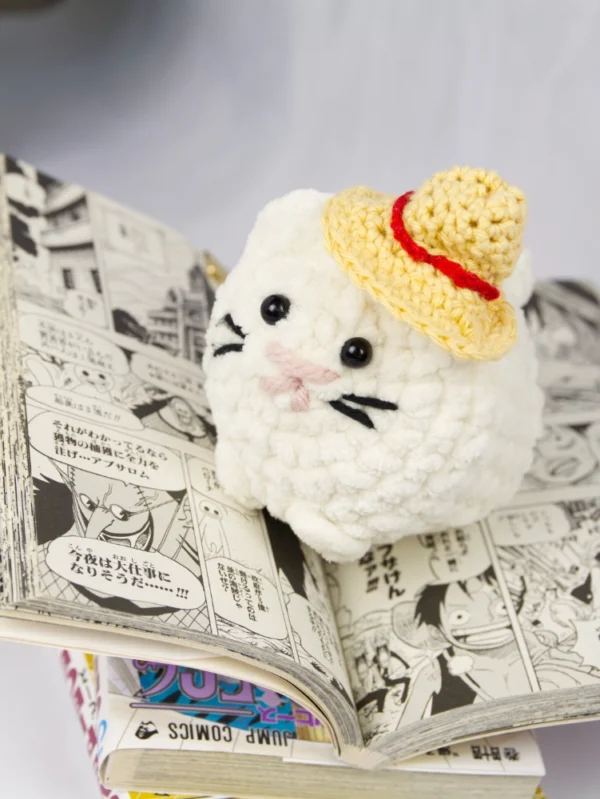 A white crochet cat with a crochet strawhat on top of its head. The cat is positioned on top of an open one piece manga book.