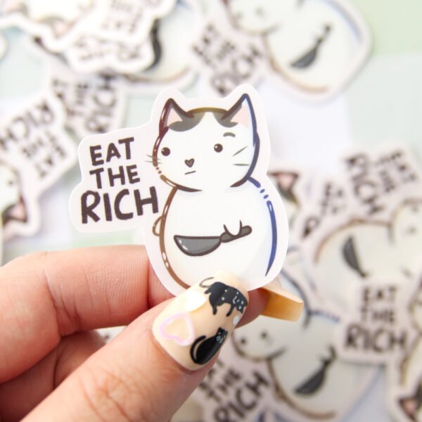 A hand holding Aa sticker of a cat holding a knife with the text eat the rich.