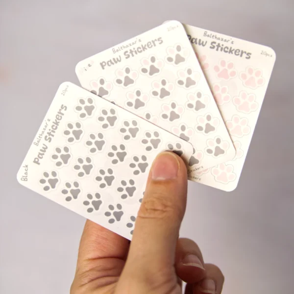a photo of the three versions of mini paw sticker sheet. a hand is holding the sheets.