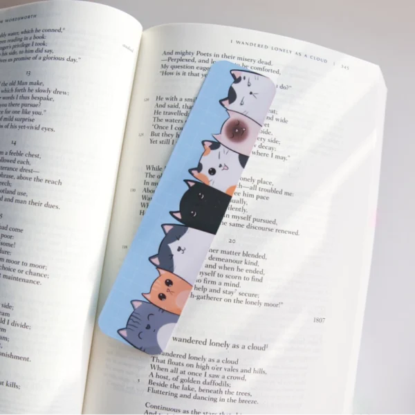 A colorful bookmark featuring a row of seven cute cat illustrations, each with unique expressions and colors, placed over an open book