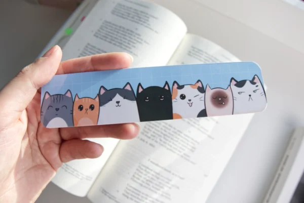 Hand holding a colorful bookmark featuring a row of seven cute cat illustrations, each with unique expressions and colors, placed over an open book