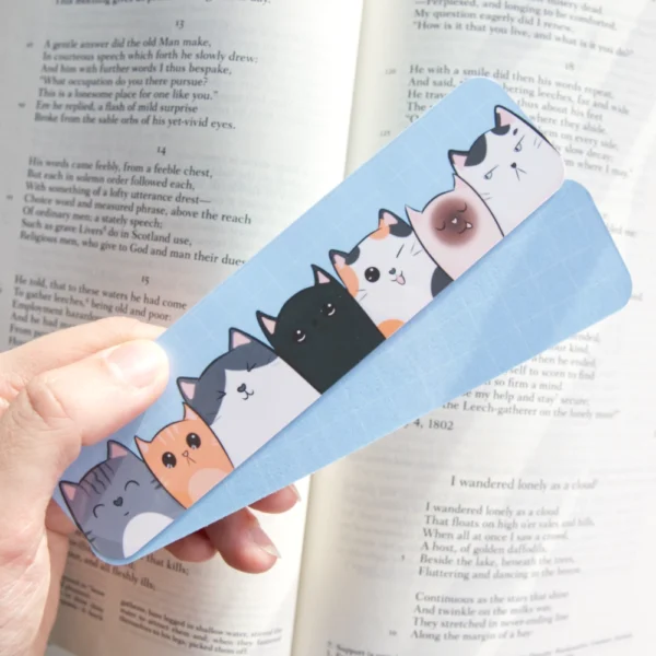 Hand holding two colorful bookmarks featuring a row of seven cute cat illustrations, each with unique expressions and colors, placed over an open book