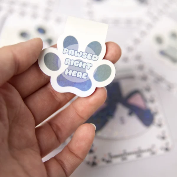 A hand holding a paw-shaped magnetic bookmark with a colorful, geometric design and the text 'Pawsed Right Here'.