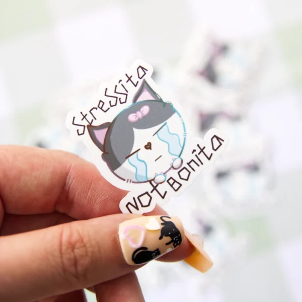 A hand holding a sticker of a crying cat with teary eyes and the phrase “stressita not bonita” written above it.