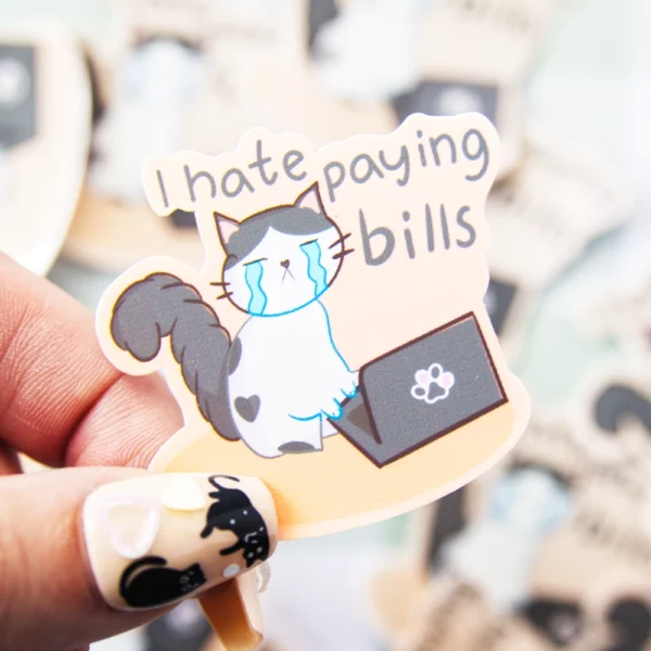 A hand holding a sticker of a cat crying in front of a computer with the words I hate paying bills over.