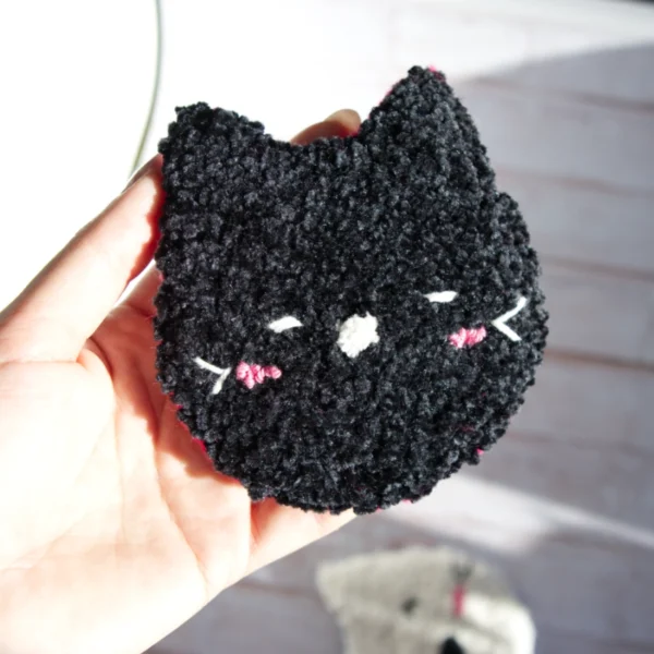 Hand holding a cat-shaped mug rug made with punch needle technique, in black, featuring playful expressions and pink accents.