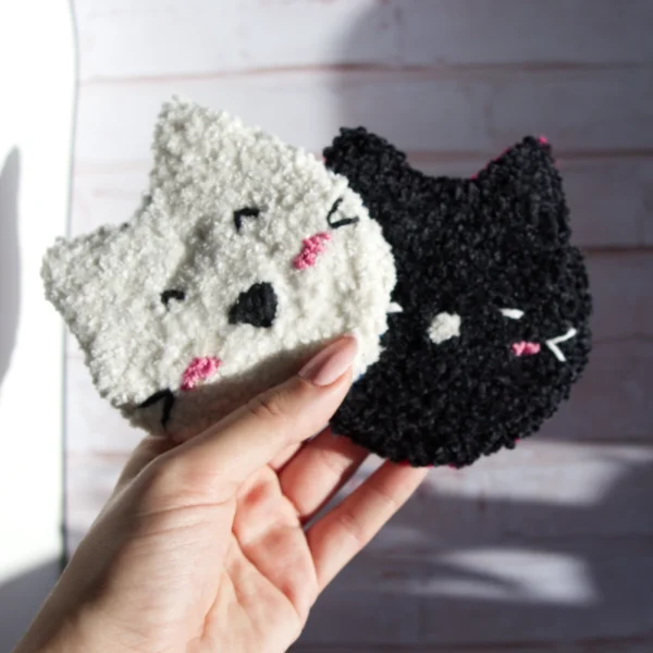 Hand holding two cat-shaped mug rugs made with punch needle technique, one in white and the other in black, featuring playful expressions and pink accents.