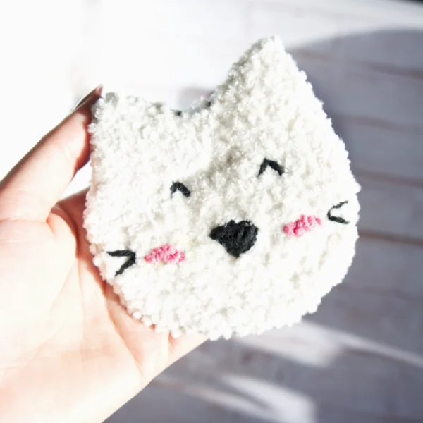 Hand holding a cat-shaped mug rug made with punch needle technique, in white, featuring playful expressions and pink accents.